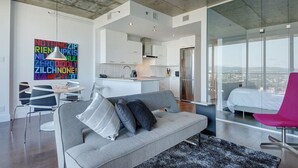 Un appartement moderne avec un canapé gris, un fauteuil rose, une cuisine et une grande fenêtre donnant sur la ville.