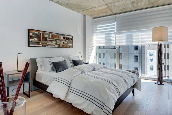 A modern bedroom with a large bed, a bedside table, a floor lamp, and a view of the city through large windows. - Les Immeubles Charlevoix - 760608 (Ville de Québec)