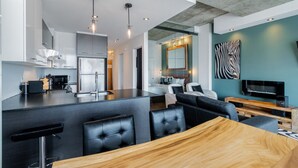 A modern kitchen with a central island, a dining area with a wooden table, and a living space with a sofa and a TV. - Les Immeubles Charlevoix-Le 760631 (Québec)