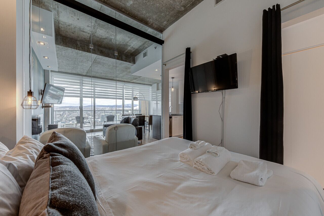 A modern hotel room with a large bed, a flat-screen TV, and a city view through large windows.