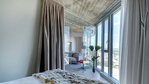 Un dormitorio moderno con una cama grande, vista de la ciudad a través de ventanales del suelo al techo y un balcón con una silla roja.