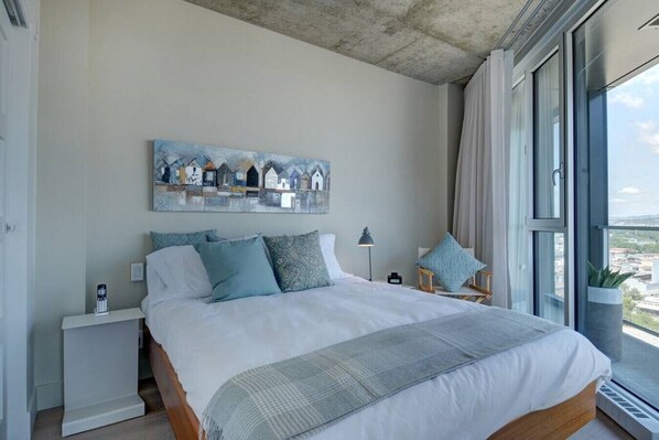 Een moderne slaapkamer met een groot bed, een nachtkastje, een lamp en uitzicht op de stad door grote ramen.