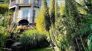 Ein großes, mehrgeschossiges Haus mit einem Balkon im Erdgeschoss, umgeben von üppigem Grün.