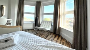 Ein Hotelzimmer mit einem großen Bett, einem weißen Sessel und Blick auf die Stadt durch große Fenster.