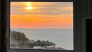 Ein Fenster mit Blick auf einen Sonnenuntergang über dem Meer, Gebäude in der Ferne und ein bewölkter Himmel.