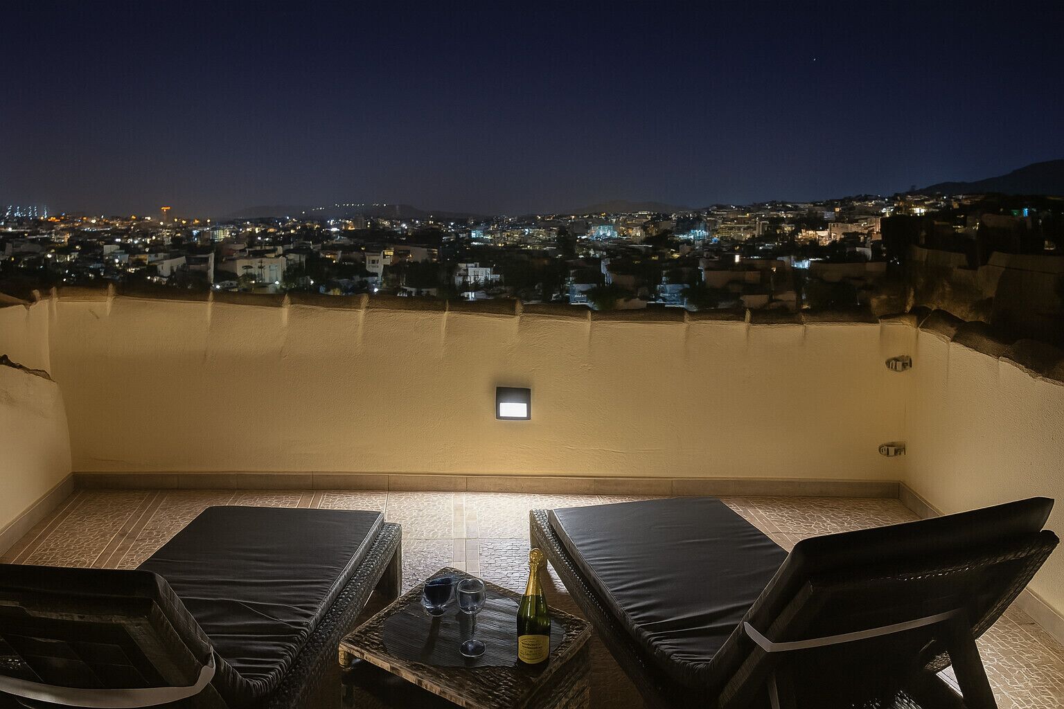 Terraço no telhado com duas cadeiras de descanso, uma mesa pequena com uma garrafa e copos, e vista para o horizonte urbano à noite.