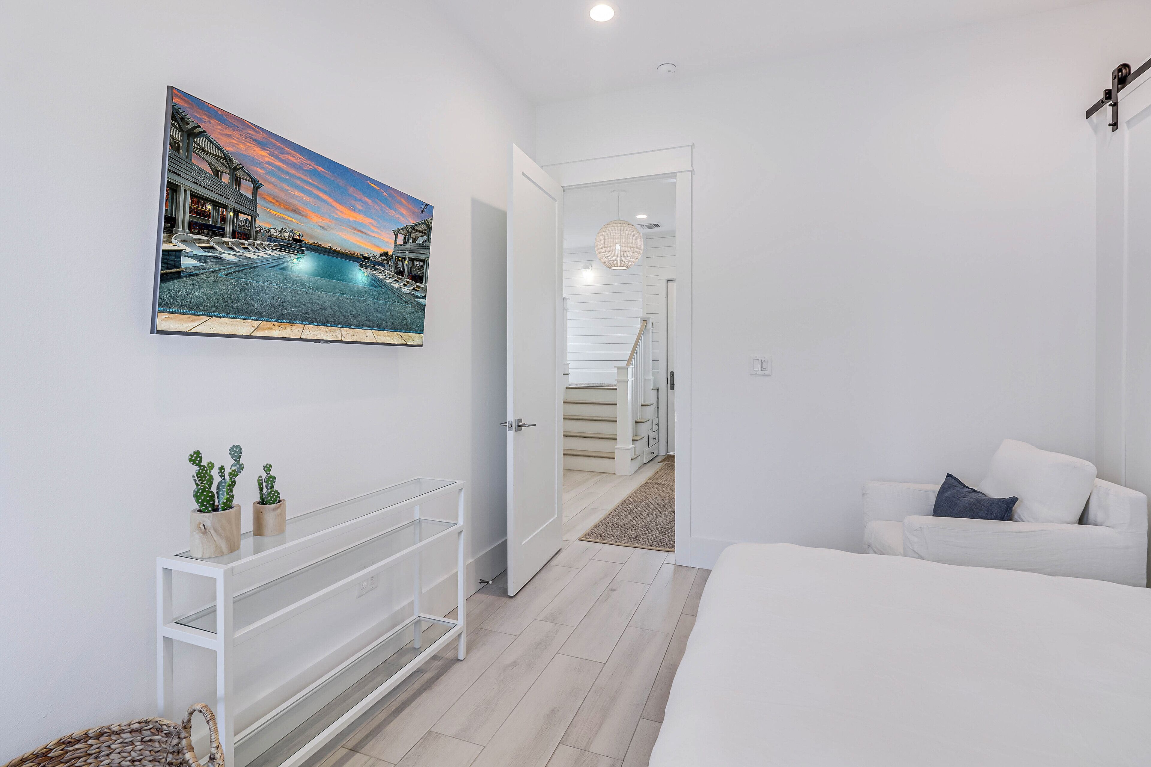 Un dormitorio moderno con una cama grande, un sofá blanco, un cuadro de una piscina al atardecer y una consola blanca con plantas.