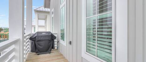 A covered wooden deck with a grill, white railings, and large windows with blinds.