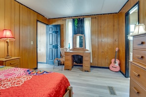 A bedroom with wooden walls, a bed with a colorful quilt, a dresser, a desk with a mirror, and a guitar.