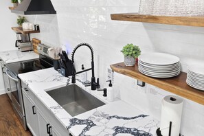 A modern kitchen with a marble countertop, a stainless steel sink, and a black faucet. There are wooden shelves above the sink holding plates and a potted plant. - Lakefront, Views, Private Dock, Boat Rental, Bunks, Near Clemson, Kayaks! (Starr)