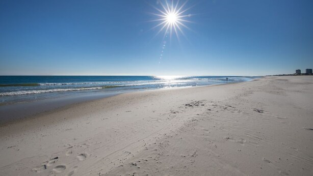 A beach with footprints leading to the water, a clear sky, and a bright sun.