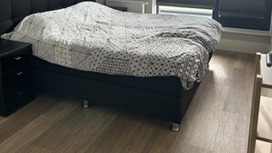 Un dormitorio con una cama grande, una mesita de noche negra, un radiador blanco y una ventana con persianas.