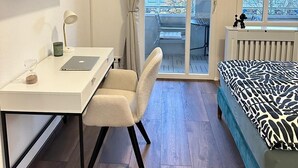 Une chambre avec un lit, un bureau avec un ordinateur portable, une chaise, un téléviseur fixé au mur et un balcon donnant sur des immeubles.