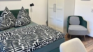 Une chambre avec un lit de couleur turquoise, des coussins à motifs noir et blanc, un fauteuil vert, une armoire blanche et une horloge noire accrochée au mur.