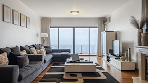 Ein modernes Wohnzimmer mit grauem Sofa, Couchtisch und Fernseher. Ein großes Fenster bietet Blick auf die Stadt.