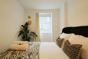 Relax in a cozy retreat with a neatly made bed, a potted plant, and a window with a curtain.