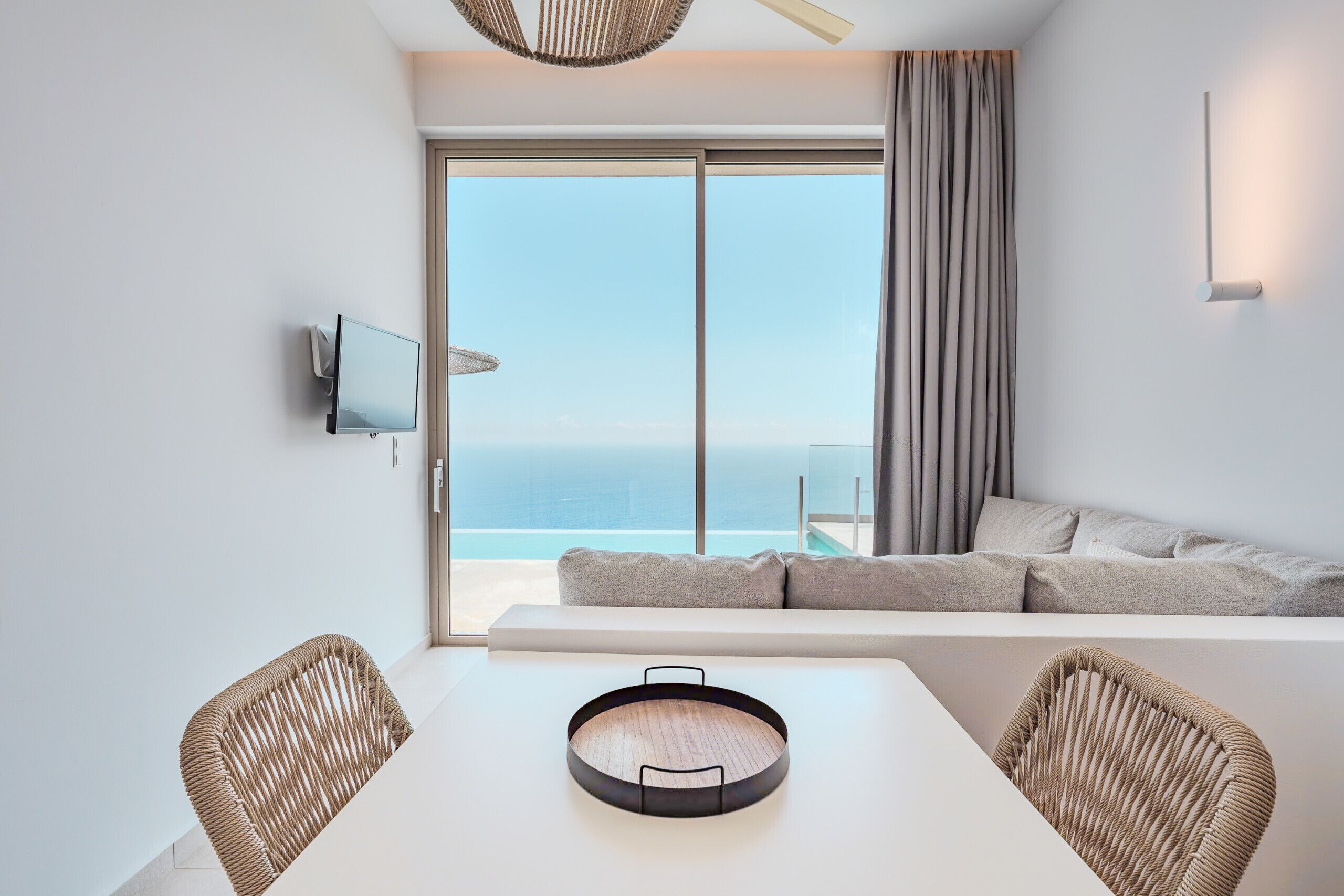 A modern room with a large sliding glass door leading to a balcony, a white table with a tray, two wicker chairs, a sofa, and a mounted television.