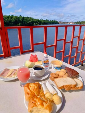 Mesa com itens de café da manhã, incluindo pão, doces, melancia e bebidas, com uma vista panorâmica da orla.