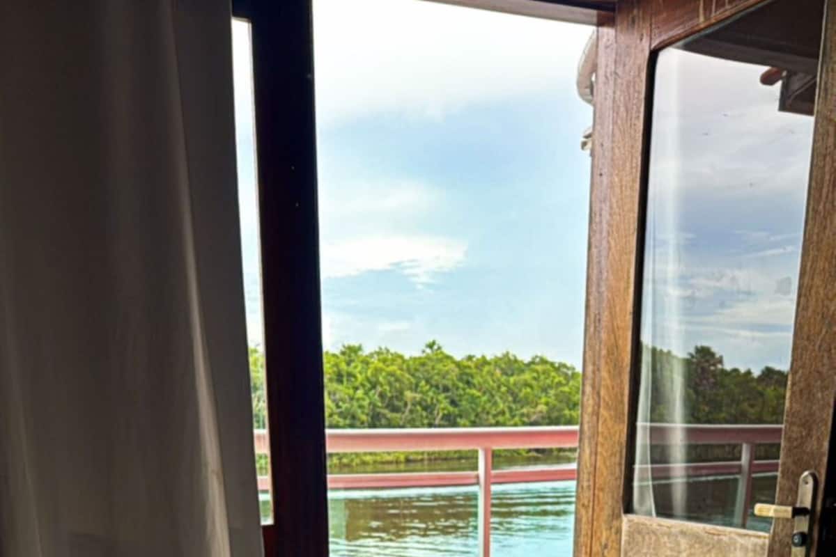 Une chambre d’hôtel avec un lit, une serviette roulée et une vue sur une piscine et de la verdure, le tout visible à travers une porte ouverte.