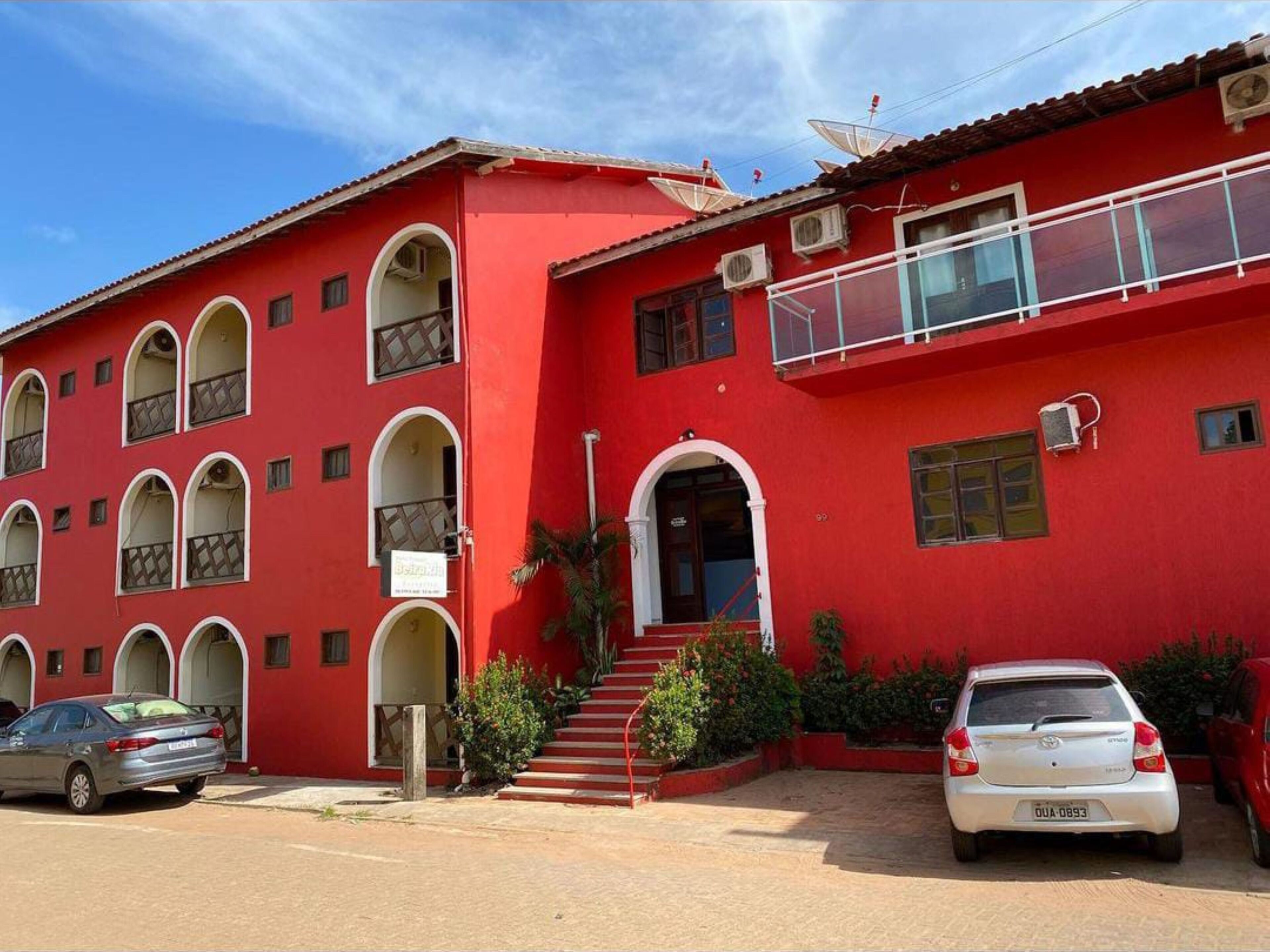 Un edificio rojo con varios balcones, una escalera que lleva a la entrada y coches estacionados adelante.