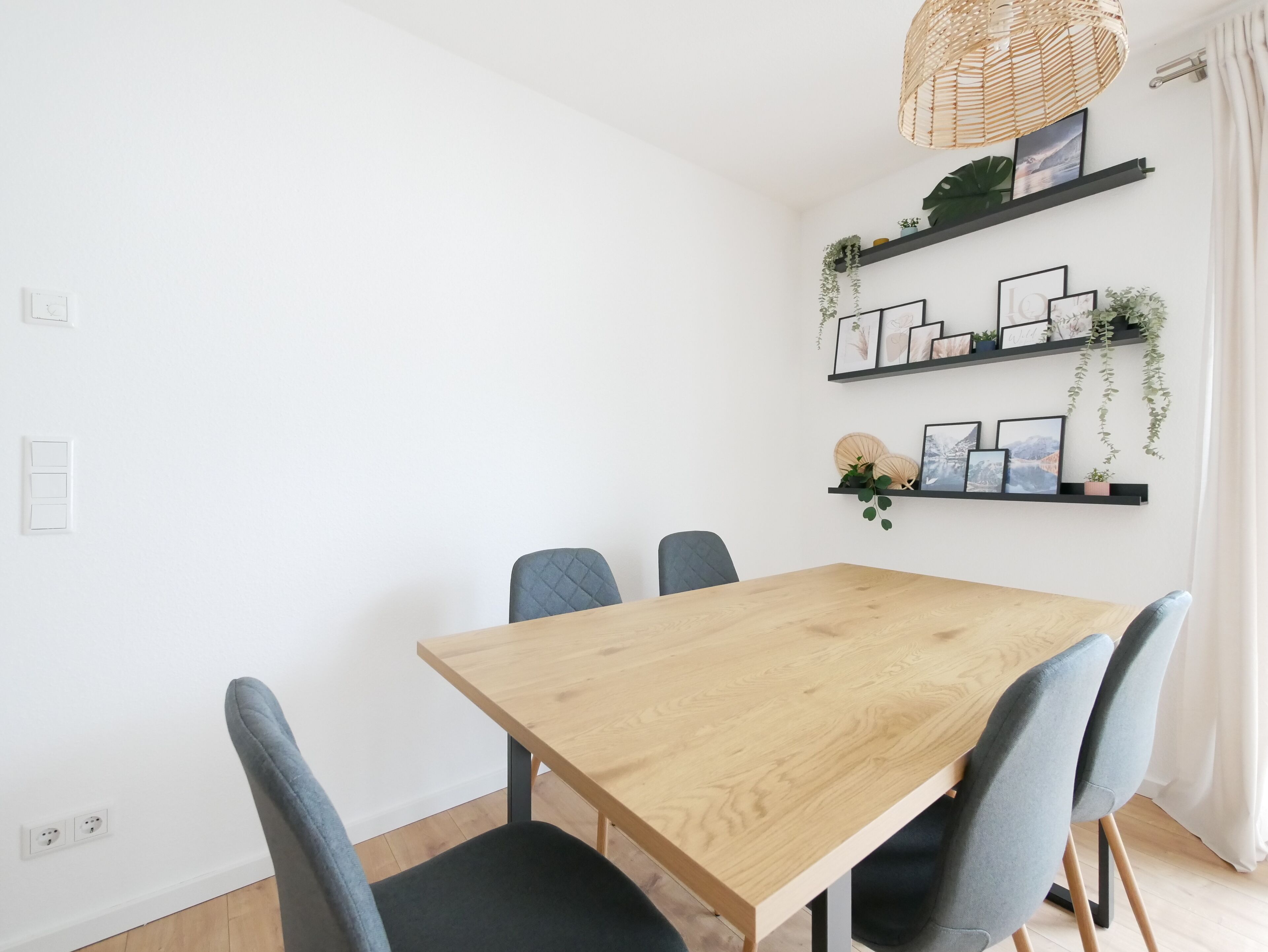 A dining area with a wooden table and four chairs, a hanging wicker light fixture, and two black shelves with framed pictures and plants.