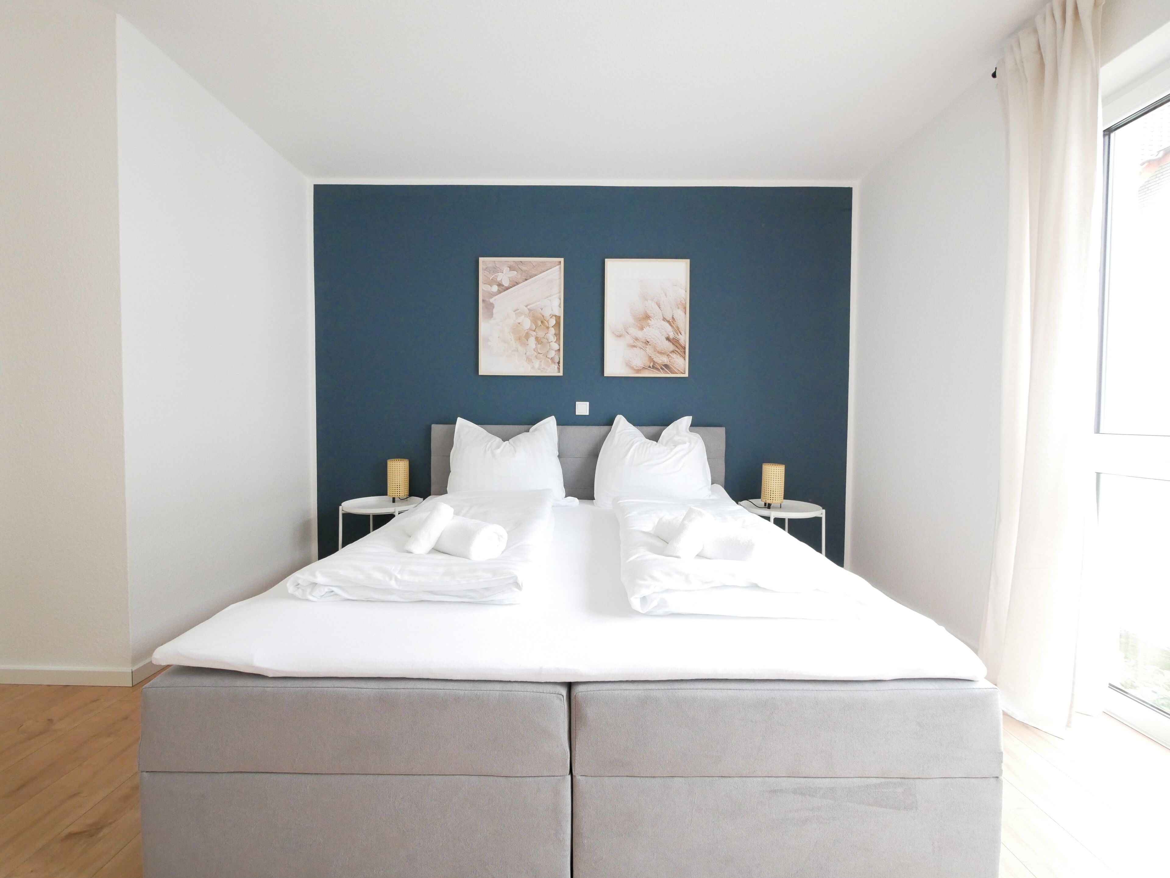 A neatly made bed with white linens and pillows, two bedside tables with lamps, and two wall art pieces above the bed.