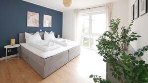 Quarto moderno com cama cinza, roupa de cama branca, parede de destaque azul, uma janela grande e uma planta em vaso.
