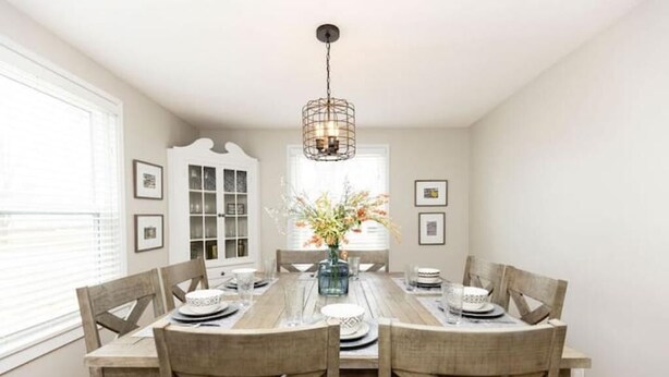 A dining room with a wooden table set for six, surrounded by wooden chairs. A chandelier hangs from the ceiling, and a vase with flowers is on the table.