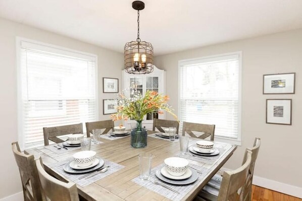 A dining room with a wooden table set for six, surrounded by chairs. A chandelier hangs from the ceiling, and a vase with flowers is on the table.