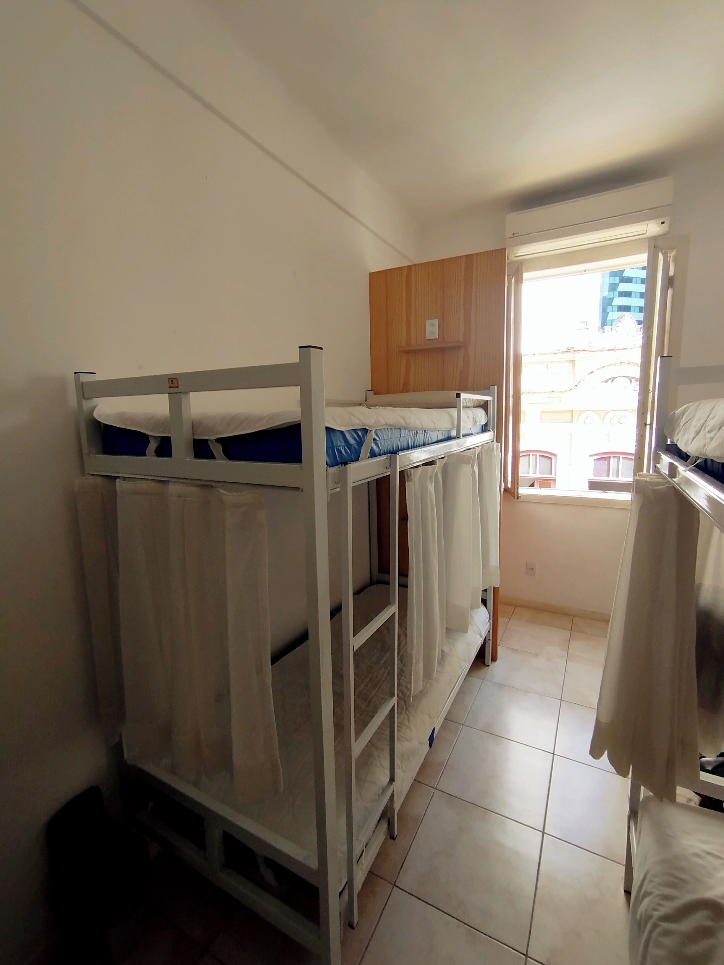 A dormitory room with bunk beds, a wooden cabinet, and a window with a view of buildings.