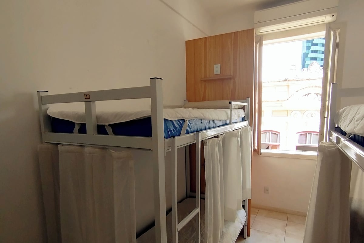 Quarto em dormitório com beliches, um armário de madeira e uma janela com vista para edifícios.