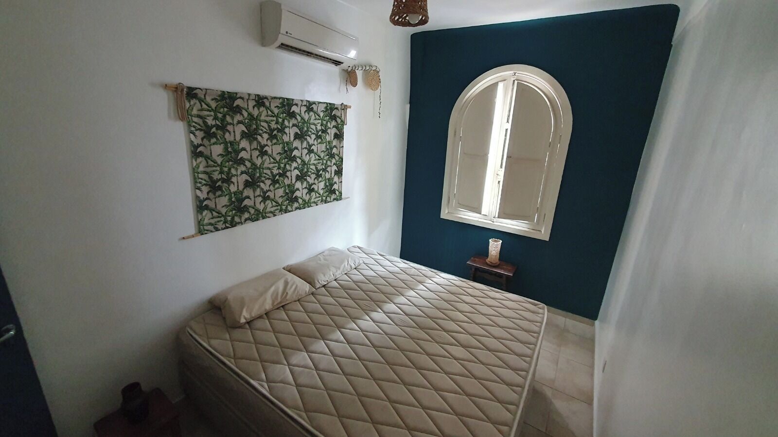 A bedroom with a bed, a wall-mounted air conditioner, a window with an arched top, and a curtain partially drawn.
