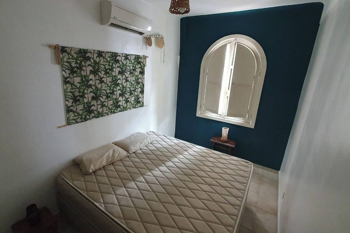 A bedroom with a bed, a wall-mounted air conditioner, a window with an arched top, and a curtain partially drawn.