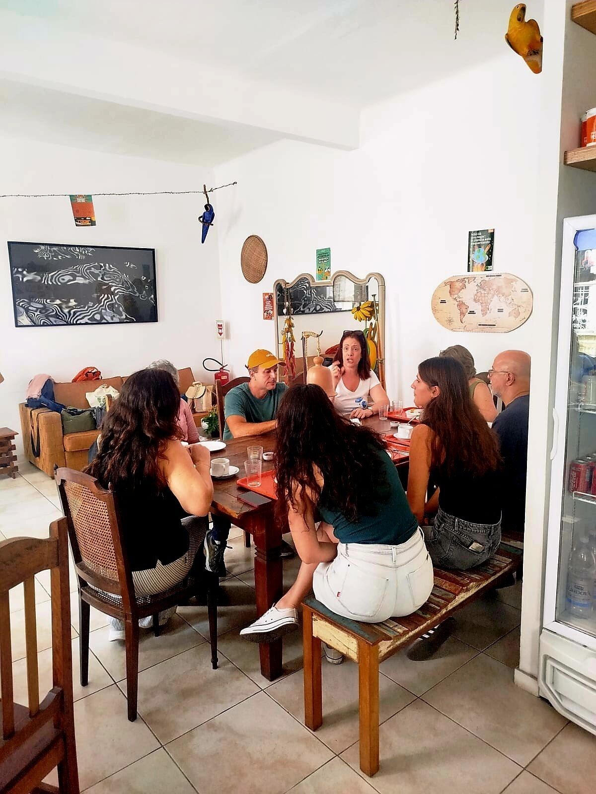 A group of people seated around a wooden dining table in a room with a tiled floor, a large mirror, and various wall decorations including a world map.