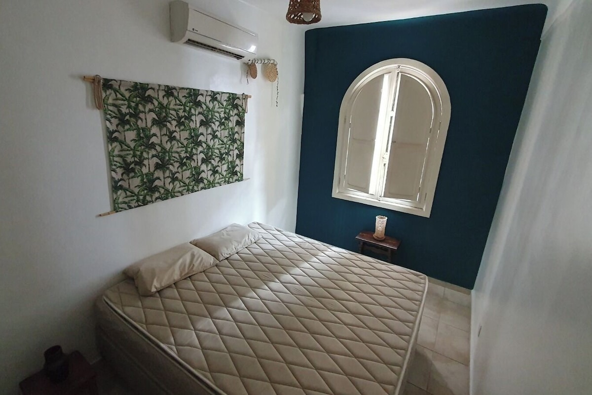 Quarto com cama, ar-condicionado fixado na parede, janela com arco e cortina entreaberta.