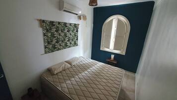 Un dormitorio con cama, aire acondicionado de pared, una ventana con arco y una cortina entreabierta.