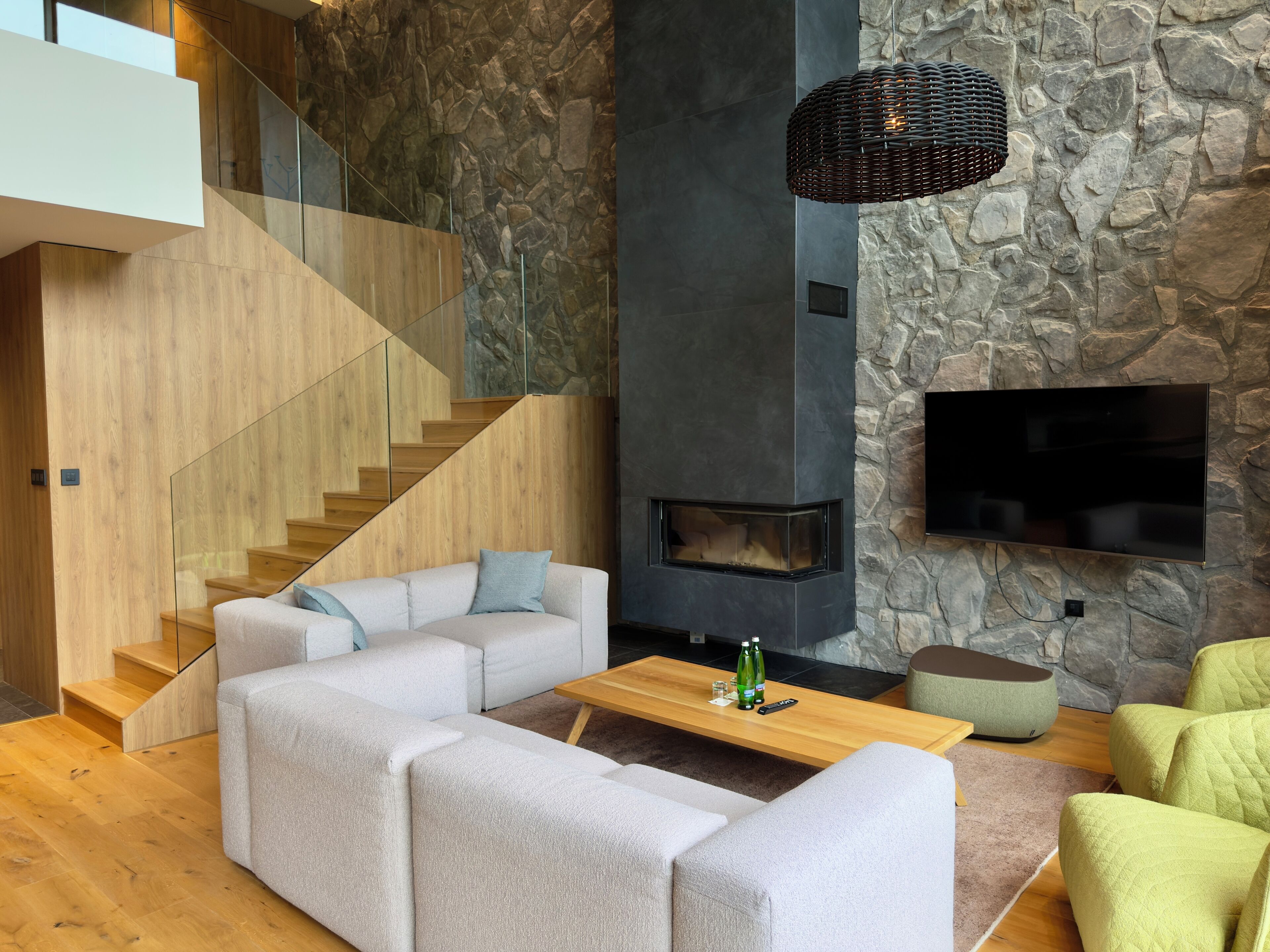 A modern living room with a stone wall, a fireplace, a flat-screen TV, and a wooden staircase.