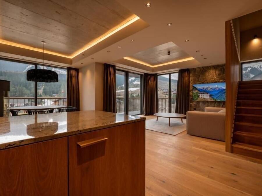 A modern kitchen with wooden cabinets, a marble countertop, and a dining area with a table and chairs. There is a large window with a view of mountains and a balcony with a sofa and a TV.