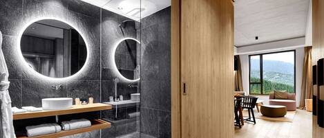 Un baño moderno con dos espejos grandes, un lavabo y una ducha. La habitación tiene una puerta de madera que da a una sala de estar con un sofá y un televisor.