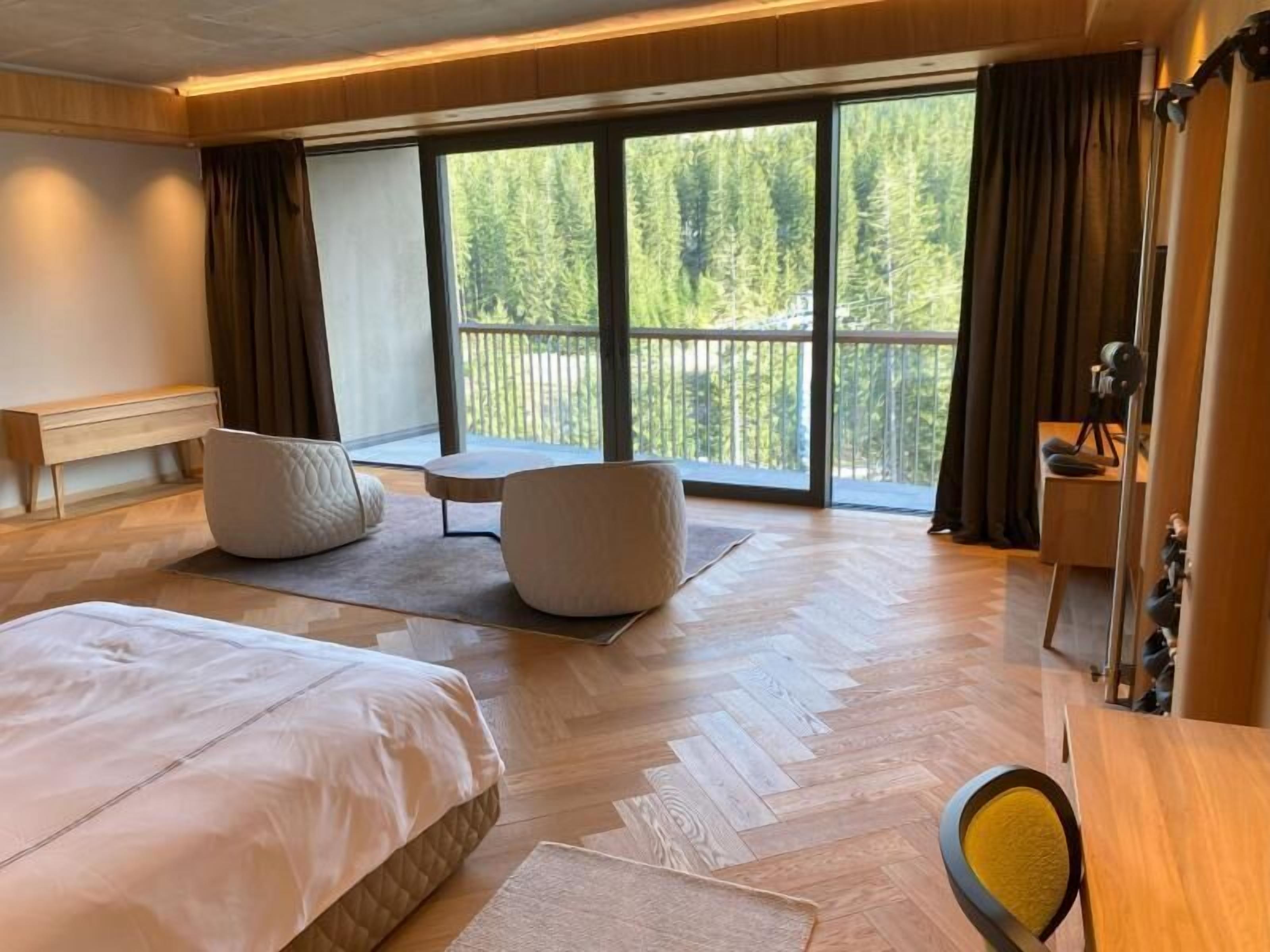 A hotel room with a large bed, two armchairs, a small table, and a balcony with a view of trees.