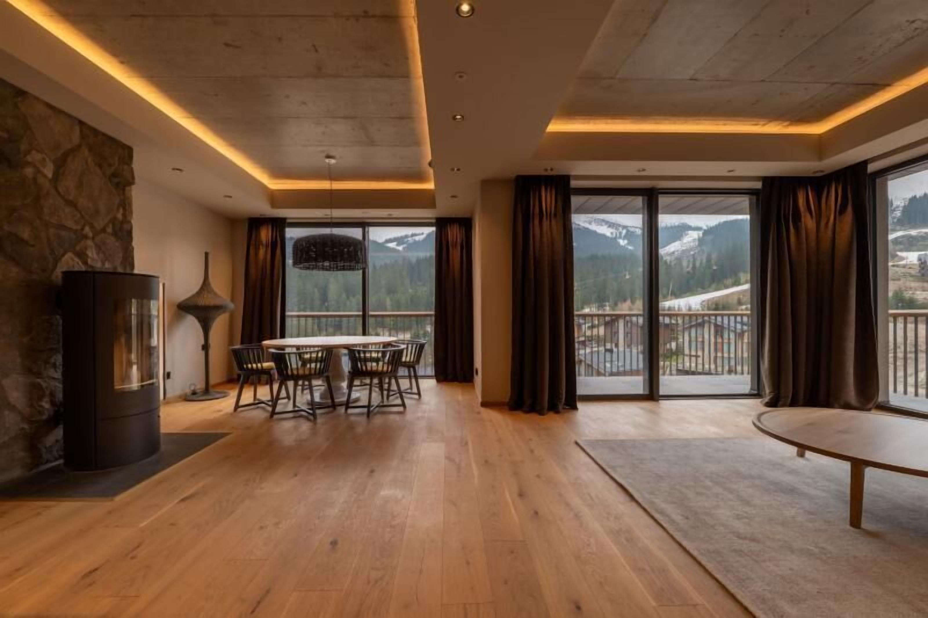 Una habitación amplia con piso de madera, una mesa redonda de comedor con sillas, pared de piedra, chimenea y ventanas grandes con cortinas.