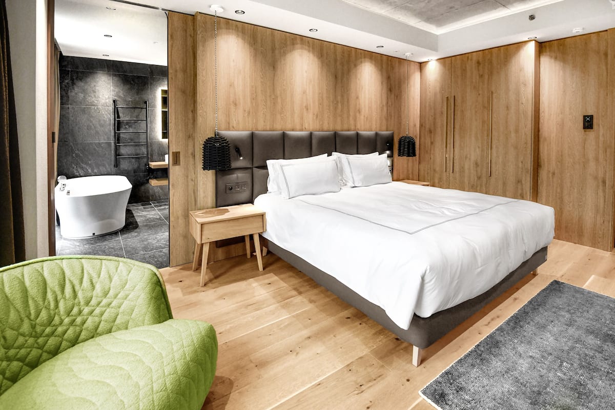 A modern bedroom with a large bed, a green chair, a wooden headboard, and a bathroom visible in the background.