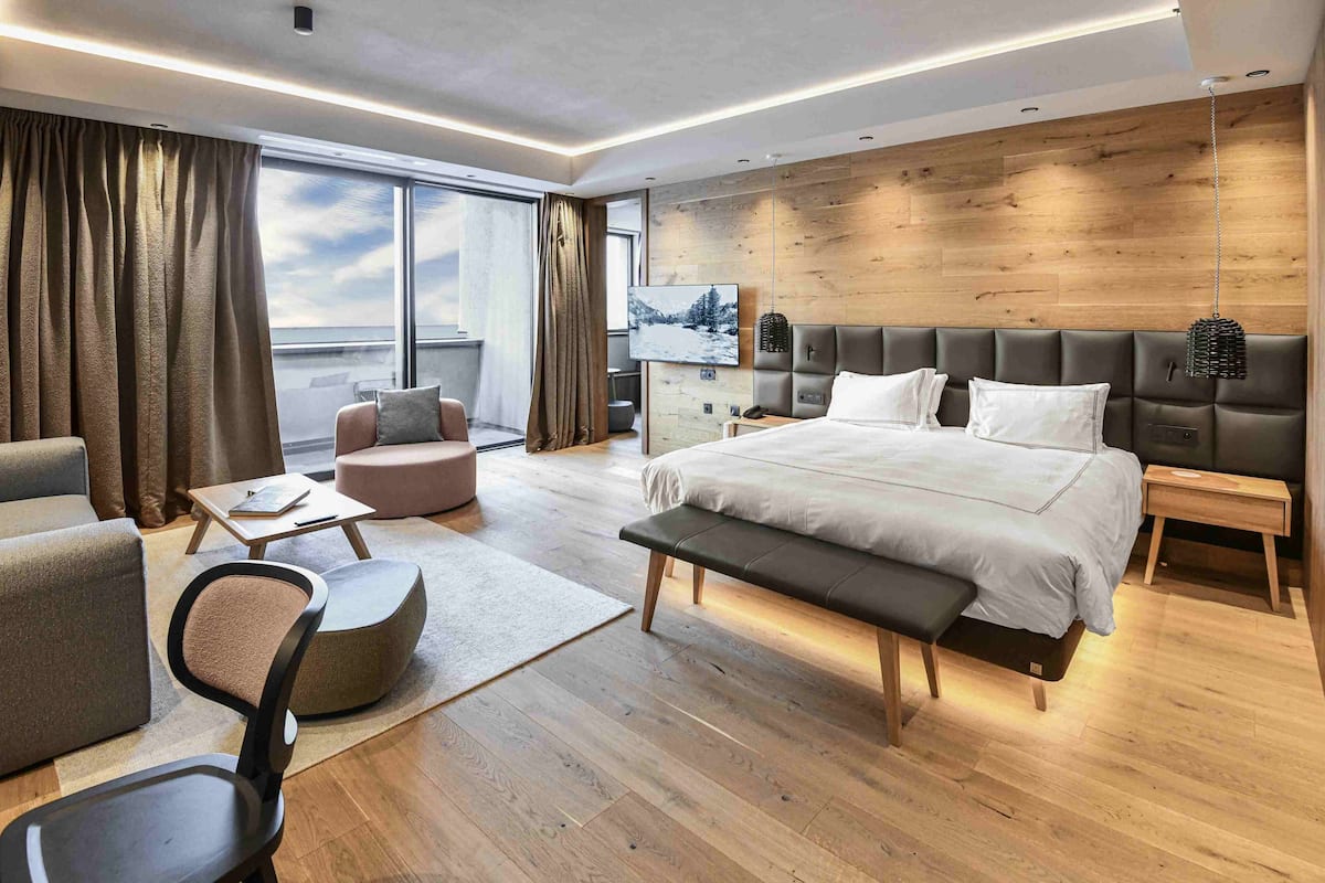 A modern hotel room with a large bed, a seating area with a bench and chairs, a small table, and a large window offering a view.