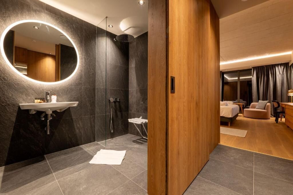 A modern bathroom with a round mirror, a sink, and a shower. The room has a wooden door and a bed in the adjacent bedroom.