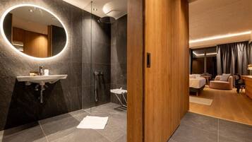 A modern bathroom with a round mirror, a sink, and a shower. The room has a wooden door and a bed in the adjacent bedroom.
