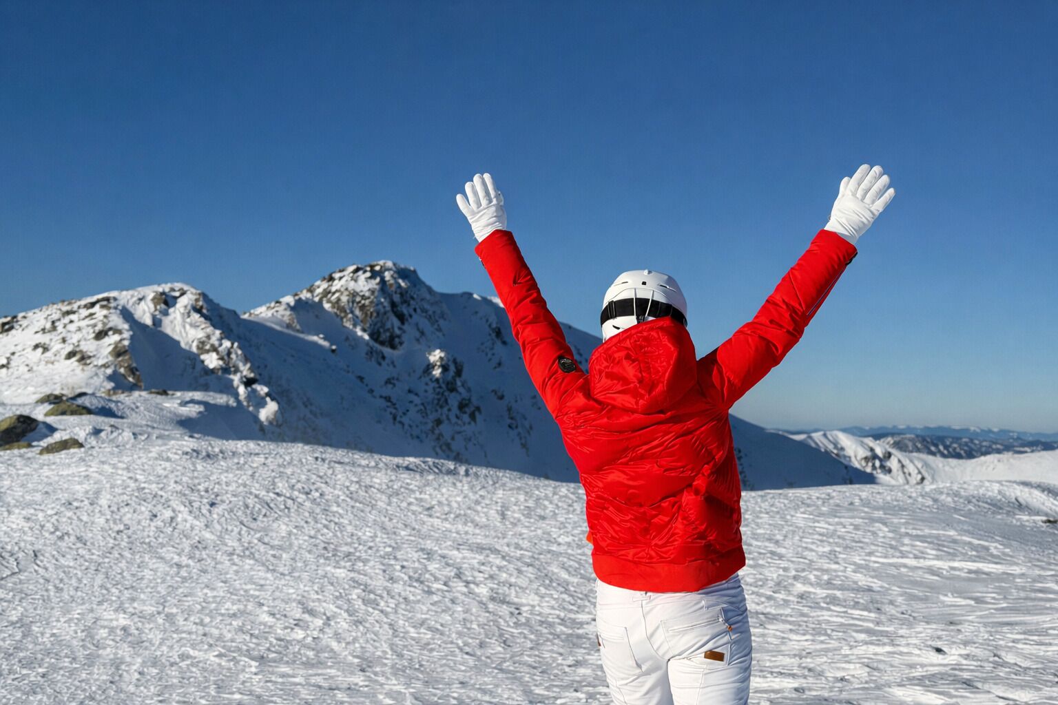 Una persona con chaqueta roja y pantalones blancos de pie en una montaña nevada, con los brazos levantados, mirando hacia un cielo azul despejado y picos cubiertos de nieve.