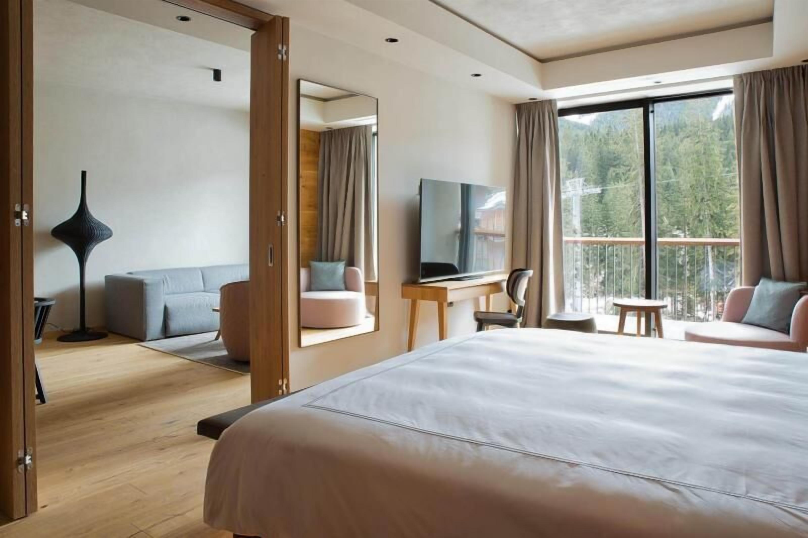A modern hotel room with a large bed, a TV, a desk, and a balcony with a view of the forest.