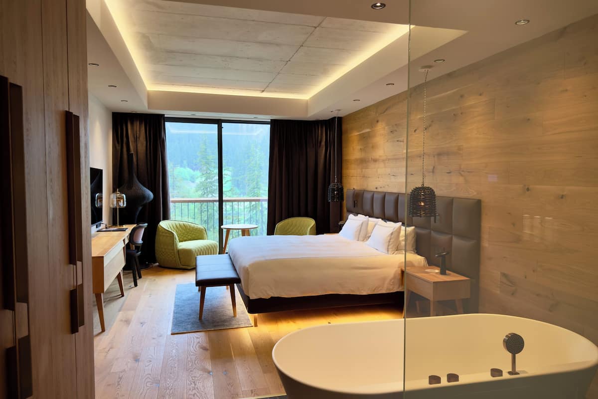 A modern hotel room with a large bed, a sitting area with a desk and chair, a glass-enclosed bathroom with a bathtub, and a view of trees through the window.