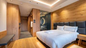 Una habitación de hotel moderna con piso de madera, una cama grande con ropa blanca, un banco y una mesita de noche.