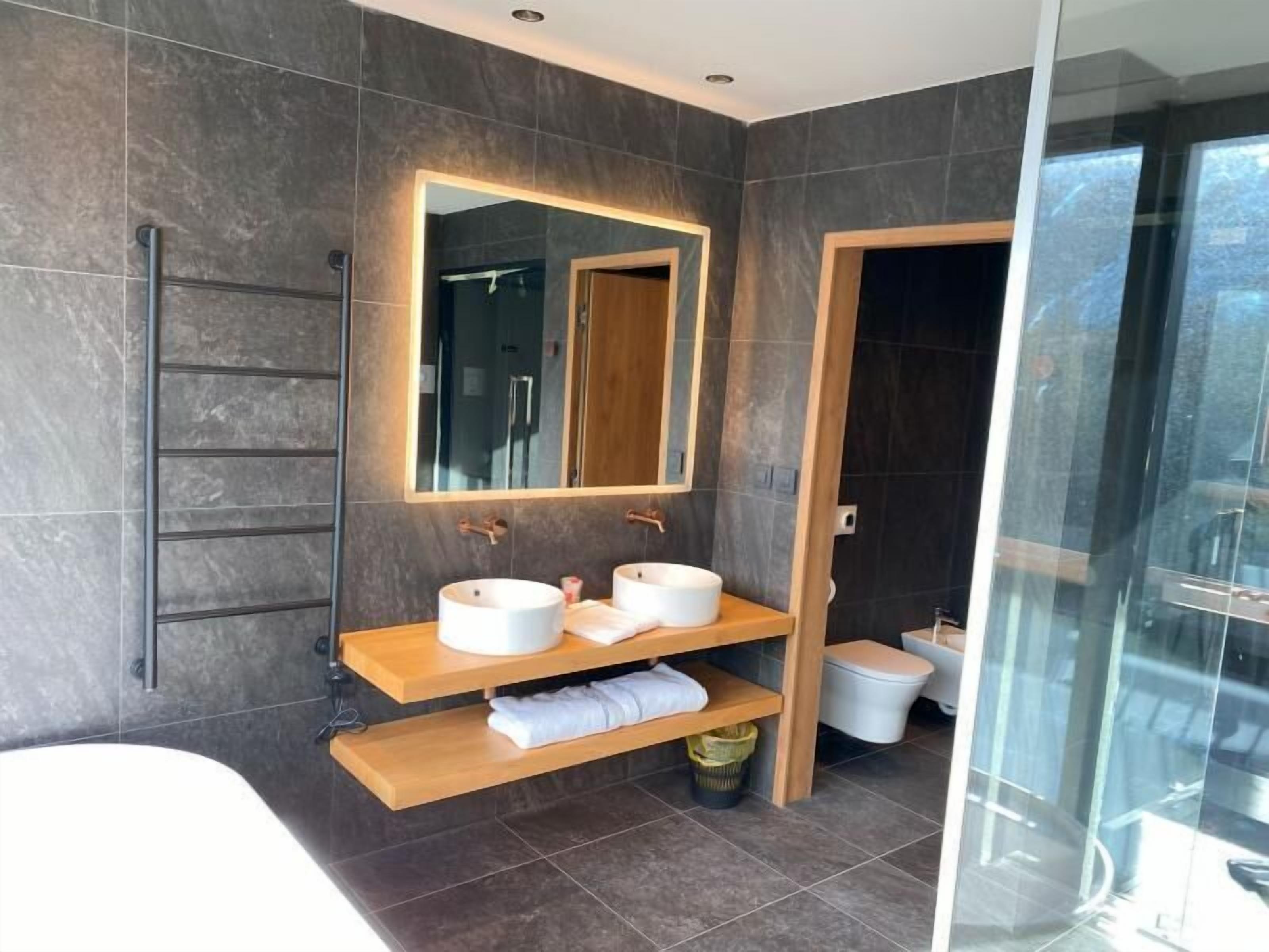 A modern bathroom with dark tiled walls, a large mirror, two sinks, a shower, and a glass-enclosed shower area.